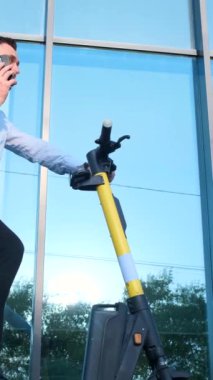 Ofis müdürü elektrikli scooter kullanıyor ve akıllı telefonla konuşuyor. Çevre dostu ulaşım. Dikey video