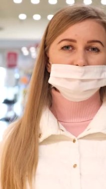 Sarışın kadın markette tıbbi maske takıyor ve Coronavirus 'a karşı korumak için kameraya bakıyor. Dikey video