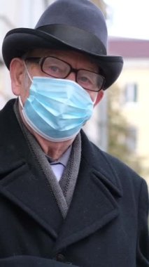 Yüzünde mavi bir maske olan yaşlı bir adamın portresi, bir şehir caddesinde dururken kameraya bakıyor. Salgın bir covid-19 hastalığının önlenmesi. Omicron virüsü. Dikey video