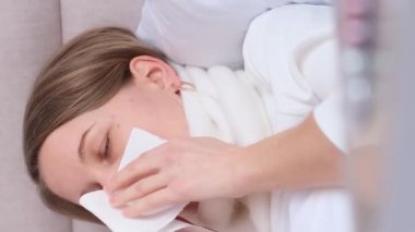Soğuk algınlığı olan Avrupalı bir kadın bandajlı bir boğazla yatakta yatıyor. Evde mevsimsel grip ve soğuk algınlığı tedavisi. Dikey video