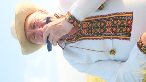 ウクライナの民族衣装の孤独な才能ある高齢者が マウスオルガン ハーモニカを演奏し フィールドの真ん中に座っています 刺繍されたシャツの祖父 バーティカルビデオ — ストック動画