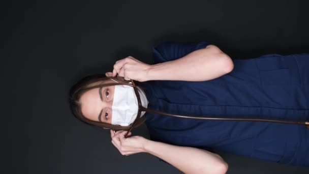 垂直图像 一个年轻女医生在黑暗背景下的画像 戴着医疗面罩的医生把听诊器放在管子上 治疗师 儿科医生Pediatrician 垂直录像 — 图库视频影像