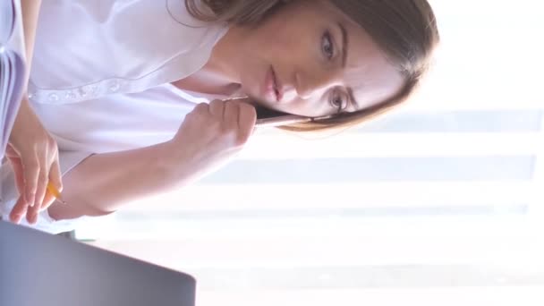 一位穿着正式西装的女商人坐在公司总部的办公桌前 一边打电话一边聊天 在笔记本电脑上工作的女商人 垂直录像 — 图库视频影像