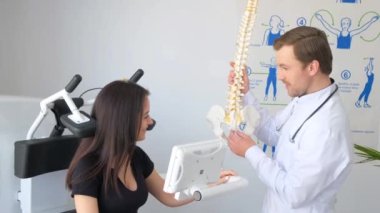 Travmatologla konsültasyonda olan esmer bir kadın. Doktor, omurganın iskelet modelini kullanan bir müşteriye danışıyor. Görsel denetim.