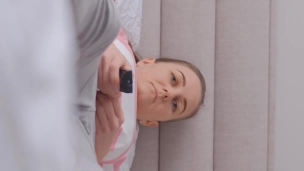 ある女性がテレビのリモコンを持っていて 寝室の広いベッドに横たわっている 疲れた女性 バーティカルビデオ — ストック動画