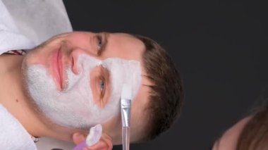 Kaplıcada yüz bakımı yaptıran genç bir adam. Bir insanın yüzünde kil maske. Bir kozmetolog yüzüne nemlendirici bir maske takarak yüz masajı yapar. Dikey video