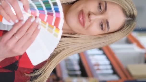 一个年轻的金发姑娘在五金店里选择彩绘和刷子 一个漂亮的女孩在油漆商店里买东西 垂直录像 — 图库视频影像