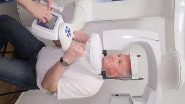 Röntgen makinesindeki yaşlı bir adamın dikey videosu. Sefalometrik soketli. Yaşlı bir adam onun yüzünün bir resmini çeker. Yüzün görüntülenmesi