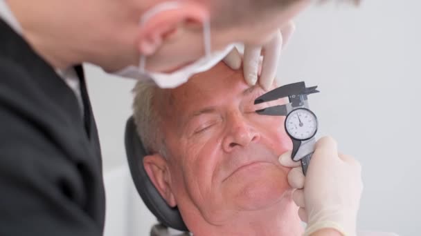 プラスチック外科医は 目の領域の賢い皮膚を検査し キャリパーでそれを測定します 高齢者の顔にプラスチック手術の準備 — ストック動画