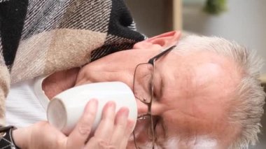 Gözlüklü büyükbaba çay içiyor, üşütmüş ve burnu akıyor. Hasta yaşlı adam. Dikey video