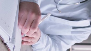 Gözlüklü ve üniformalı profesyonel genç doktor ofisinde evrak işleriyle uğraşıyor. Teşhis, tedavi planı yazıyorum. İş yerinde bir doktor. Dikey video