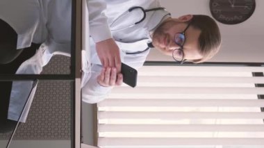 Ofisteki masada oturan genç profesyonel doktor akıllı telefona bakıyor. Doktorlar gündüzleri çalışıyor. Özel terapist muayenehanesi. Dikey video