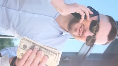 Elinde dolarlar olan genç bir patron akıllı bir telefonla konuşuyor. İşletme müdürü konsepti. Dikey video