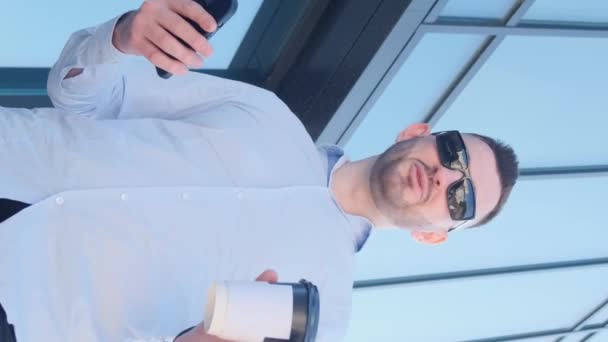 一个英俊的男人站在一座摩天大楼附近 一边喝咖啡 一边对着摄像机说话 成功的时髦商人在街上和他一起喝咖啡 垂直录像 — 图库视频影像