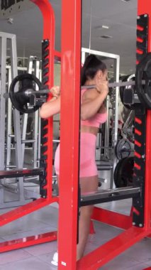 Vücut geliştirici kız omuzlarında ağır bir halter tutuyor ve çömeliyor. Bir güç kaldırıcı dayanıklılığını ve uygunluğunu geliştirir. Profesyonel sporlar. Dikey video