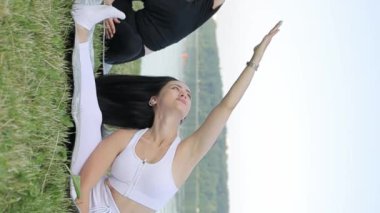 İki güzel, narin genç kadın yoga yapıyor, bir paspasın üzerinde oturuyor ve şehir parkında ellerini kaldırıyor. Güneşli bir günde dışarıda yoga yapan kadınlar. Dikey video