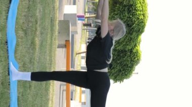 Genç bir kadın, Yogi, parkta yoga yapan bir sporcu, egzersiz yapıyor, bacağını havada tutuyor, akrobatik bir unsur, vücudunu esnetiyor. Dikey video