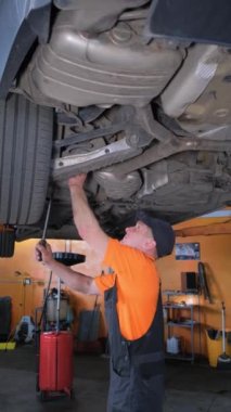 Üniformalı bir tamircinin kaldırılmış bir arabayla araba tamirhanesinde çalışmasının dikey videosu..