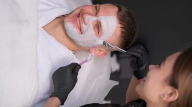 Bir güzellik uzmanına yüz maskesi takan yakışıklı, gülümseyen bir adamın dikey videosu. Güzellik salonunda bir adam.