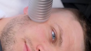 Modern elektrikli masaj aleti kullanan genç ve çekici bir adamın yüz masajı. Erkek kozmetolojisi gençleştirici. Dikey video