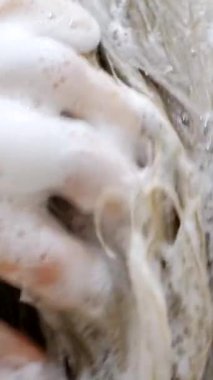 Genç bir kadın saçını şampuanla yıkar ve ellerini köpükle kaplar. Bir kadın saçını duşta şampuanla yıkar. Dikey video