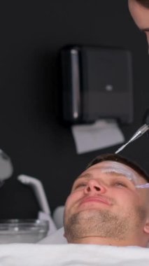 Eldiven takan bir dermatolog bir adamın yüzüne maske takar. Dermatoloji kliniğinde bir adamın yüzüne cilt bakımı. Terapötik yüz maskesi. Dikey video