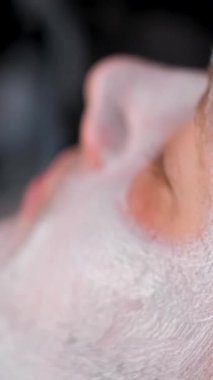 Adamın yüzüne tıbbi maske takan bir adamın yüzünü yakından çek. Bir cildiyeci, bir fırçayla yenileyici bir maske takar. Dikey video