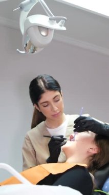 Dikey video. Genç bir dişçi modern bir diş muayenehanesinde oturan genç bir hastanın dişlerini tedavi ediyor. Modern diş teknolojileri. Dikey video