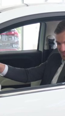 Bir adam bir galeride yeni bir elektrikli arabada oturuyor ve yeni bir araba satın alınmasını kutluyor. Bir araba al. Araba satış konsepti. Dikey video