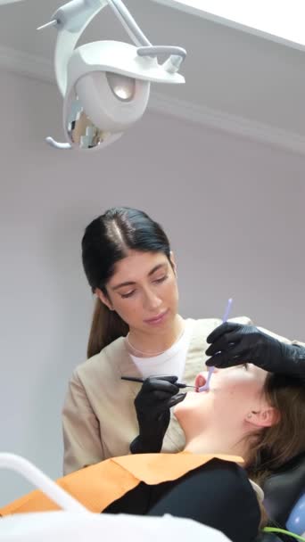 バーティカルビデオ 若い女性歯科医は 現代の歯科オフィスに座っている若い患者の歯を治療します 現代歯科技術について バーティカルビデオ — ストック動画