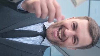 Zengin bir adam dolarları saçıyor ve kameraya gülümsüyor. İş ve finans kavramı. Zengin bir adam sevinir. Dikey video