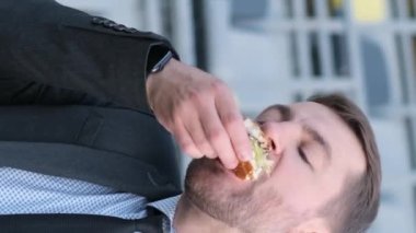 Genç bir Amerikalı ofis çalışanı dışarıda öğle yemeği yiyor, bir ofis binasının yanındaki bankta otururken sandviç yiyor. Dikey video