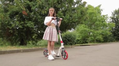 Şehir parkında bir kız scooter kullanıyor. Çocuk mutlu, doğası güzel, güneşli bir gün. Çocukların aktif şehir eğlencesi.