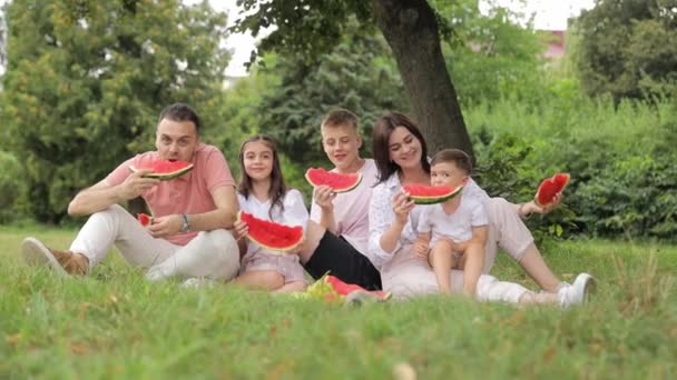 ヨーロッパの幸せな家族はピクニックでスイカを食べ 緑の庭で休息を楽しんでいます 彼らは微笑み 赤いジューシーなフルーツを味わっています サマータイム — ストック動画