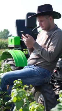 Genç bir çiftçinin portresi traktörün yanındaki tarlada dinleniyor ve bir alet kullanıyor. Ekoloji kavramı, ulaşım, çiftçiler, temiz hava. Dikey video
