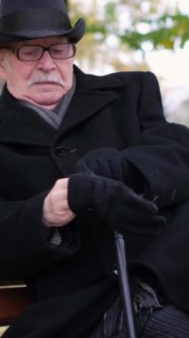 Siyah elbiseli yaşlı bir adam bir bankta oturur ve siyah eldivenler giyer. Sonbahar parkında dinlen. Dikey video