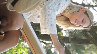 Sarışın bir kız yazın açık havada tuvale resim yapar. Parkta güneşin altında durur. Dikey video.