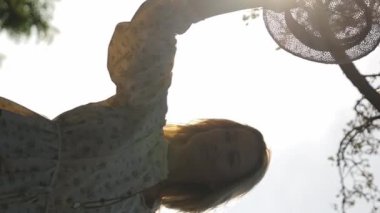 Gün batımında şapka takan ve kameraya bakıp gülümseyen güzel bir kızın silueti. Güneşli bir günde poz veren genç bir kadın. Dikey video.