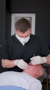 Dikey video, erkek bir dişçi yaşlı bir büyükbabanın dişlerini tedavi ediyor. Diş tedavisi prosedürü. Bir dişçinin işi. Dikey video.