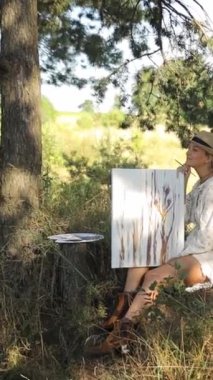 Bir kız sanatçı çam ormanında bir resim çiziyor. Açık havada güzel bir sanatçı kız. Arkaplan manzarası ve yazın ormanda yaratıcılık yaratır. Dikey video.