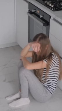Adet sancısı çeken yorgun bir kadın, mutfak odasında yerde oturuyor. Adet döngüsü. Dikey video.
