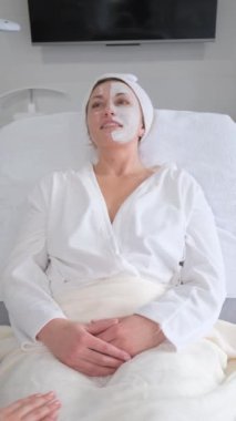 Vertical video. Relaxed middle-aged woman lying in a beauty salon with a mask on her face. Anti-aging care.