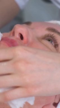 Güzellik uzmanı, kozmetikçi bir kadının yüzünü maske sonrası peçetelerle siler. Yüz için yenileme prosedürleri. Yüzün güzelliğine dikkat et. Dikey video