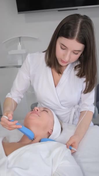 垂直录像 美容师用电动按摩器在美容院给一位中年妇女按摩脸 中年妇女面部皮肤的再生 垂直录像 — 图库视频影像