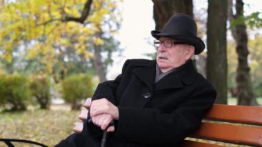 Bir sonbahar parkındaki bankta oturan şapkalı ve gözlüklü yaşlı bir büyükbaba. Yaşlı bir insandaki depresyon durumu.