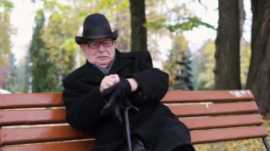 Gri bıyıklı büyükbaba sonbahar parkında bir bankta otururken eldiven takar. Serin sonbahar havası.