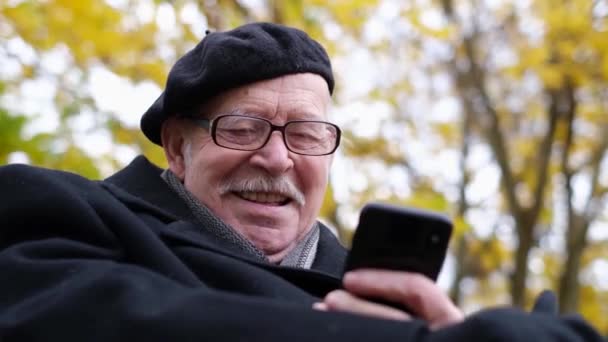 Staruszek Siedzi Ławce Parku Patrzy Komórkę Się Śmieje Używanie Gadżetów — Wideo stockowe