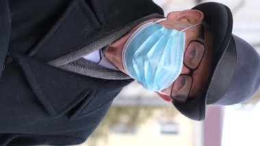 Yüzünde mavi bir maske olan yaşlı bir adamın portresi, bir şehir caddesinde dururken kameraya bakıyor. Salgın bir covid-19 hastalığının önlenmesi. Omicron virüsü. Dikey video