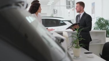 Genç bir aile araba galerisinden yeni bir araba alır, takım elbiseli bir erkek satıcı ile tezgahta durur, belgeleri doldurur ve konuşur. Genç bir çift bir araba galerisinde satış elemanıyla konuşuyor..