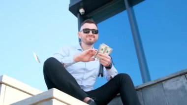 Zengin bir genç adam merdivenlerde oturur, gülümser ve dolarları saçar. 100 dolarlık banknotları olan mutlu bir işadamı..